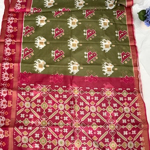 May include: An olive green saree featuring a repeating pattern of white and pink elephant motifs. The fabric includes a wide magenta border with intricate geometric designs and a matching magenta end piece with detailed diamond-shaped patterns.