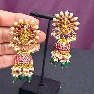 May include: Gold-toned earrings with a sunburst design, featuring a central figure, red and white accents, and dangling pearls and green beads. The earrings have a bell-shaped bottom.