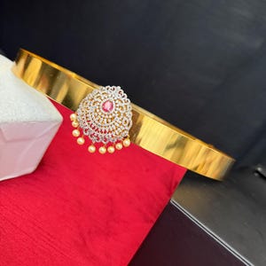 May include: A gold-toned belt with a circular, ornate centerpiece. The centerpiece features a central red stone surrounded by clear stones and dangling pearls. The belt is displayed on a red surface.
