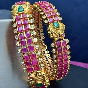 May include: Two gold bangles with a vibrant design. Each bangle features a grid of square, magenta-colored stones, accented with small, clear crystals. Green teardrop-shaped stones and ornate gold detailing complete the look. The bangles are displayed against a dark blue background.