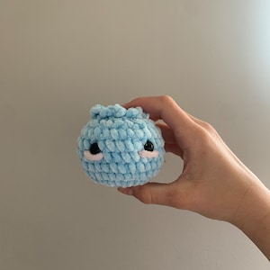 May include: A small, light blue crocheted stuffed animal with black eyes and pink cheeks. The animal is shaped like a round ball with a slightly pointed top.