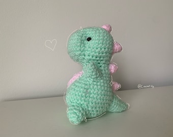 May include: A crocheted dinosaur in a light green color with pink accents on its back and head. The handmade toy has a cute, friendly appearance, perfect for children or as a decorative item. The dinosaur is sitting on a white surface.