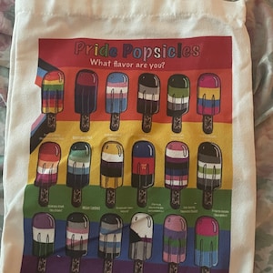 May include: A white canvas tote bag with a colorful graphic of popsicles representing different pride flags. The text "Pride Popsicles" and "What flavor are you?" is printed on the bag.