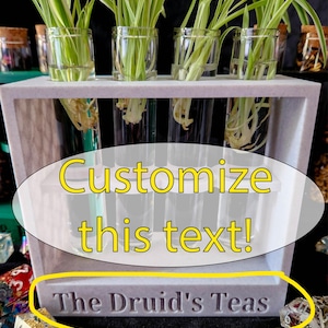 May include: A gray rectangular planter with clear glass tubes holding green plants. The text "Customize this text!" is in a yellow oval. The text "The Druid's Teas" is carved into the bottom of the planter.