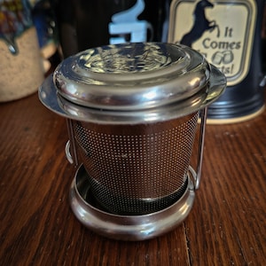 Op de afbeelding: Een roestvrijstalen thee-ei met een deksel en een gaasfilter. De thee-ei staat op een houten oppervlak.