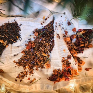 May include: An overhead shot displays three distinct piles of loose leaf tea on a white cloth. The tea varieties showcase a range of colors, including dark brown, reddish-brown, and hints of yellow and blue, suggesting a selection of herbal and black teas.