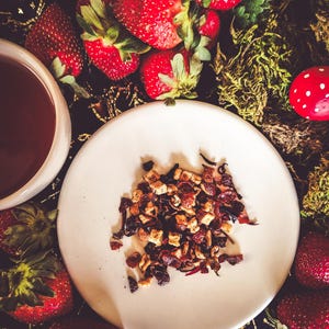 Könnte beinhalten: Ein weißer Keramikteller mit einem kleinen Haufen getrockneter Früchte und Blütentee. Der Teller ist von frischen Erdbeeren auf einem dunklen Hintergrund umgeben.