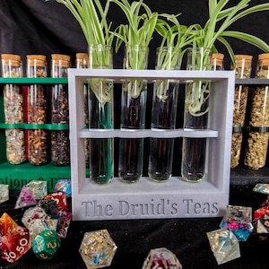 May include: A display of glass vials with plants and various dried herbs in glass jars with cork lids. The vials are in a gray wooden holder with the text "The Druid's Teas". Colorful polyhedral dice are scattered in the foreground.