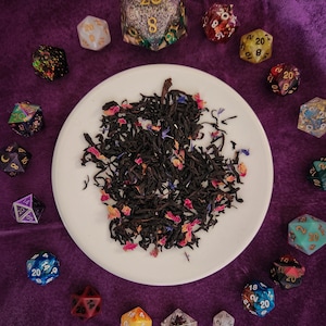 May include: A white ceramic plate with a pile of loose leaf black tea with dried rose petals and blue cornflowers. The plate is surrounded by 20-sided dice in various colors and patterns.