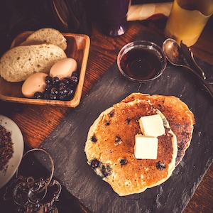 Puede incluir: Una escena de desayuno rústico con panqueques de arándanos con mantequilla, servidos sobre una pizarra negra. Acompañado de pan, huevos, arándanos, jarabe y una bebida morada en un vaso decorativo. Llaves y monedas antiguas.
