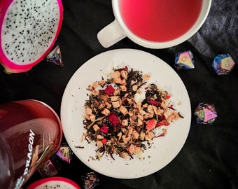 May include: A white plate with a variety of loose leaf tea, including dried fruit pieces, herbs, and flowers. The tea is surrounded by a pink teacup, two dragon fruit halves, and several iridescent dice.
