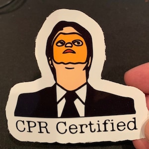 Könnte beinhalten: Ein Aufkleber mit einem Mann im Anzug, der eine gelbe Maske über dem Gesicht trägt. Der Text "CPR Certified" befindet sich unter dem Bild.