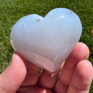 May include: A heart-shaped, pale blue crystal stone. The polished stone has small brown inclusions and is held in a hand against a backdrop of green grass. The heart shape is smooth and rounded.