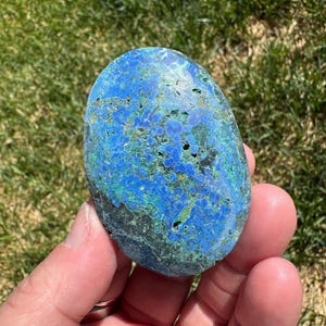 May include: A hand holding a polished, oval-shaped stone. The stone features a vibrant blue and green color palette with a textured surface. The stone's unique pattern and color variations are visible.