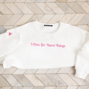 May include: A white sweatshirt with the phrase "I Can Do Hard Things" in pink script across the front. A small pink heart is on the left sleeve. The sweatshirt is displayed on a light-colored, patterned surface.