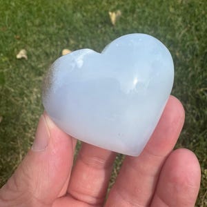 May include: A heart-shaped, light blue selenite crystal. The crystal is smooth and polished, with a slight translucence.