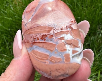 May include: A polished, oval-shaped stone with a mix of colors. The stone features a blend of reddish-brown, white, and gray hues, with a marbled appearance. The stone is held in a hand against a backdrop of green grass.