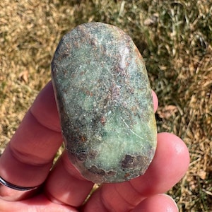 May include: A polished, oblong stone with a mottled green and brown surface. The stone is held in a hand, showcasing its smooth texture and natural variations. The stone's colors range from deep green to earthy brown.