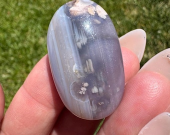 May include: A smooth, polished, gray and white agate stone with white and brown inclusions. The stone is oval shaped and has a glossy finish.