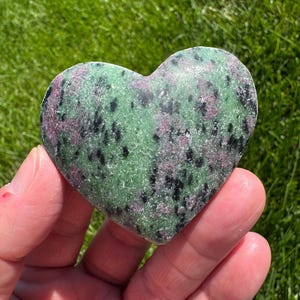 May include: A heart-shaped stone with a green base, speckled with black and pink inclusions. The polished surface of the stone is held in a hand, with a green grassy background.