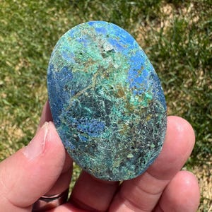 May include: A polished, oval-shaped stone with a mix of blue, green, and brown hues. The stone's surface has a textured appearance with intricate patterns and veins. The colors blend together, creating a unique and natural aesthetic.