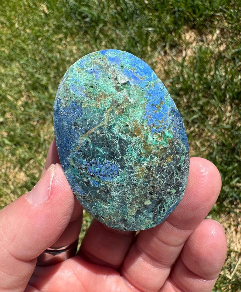 May include: A polished, oval-shaped stone with a mix of blue, green, and brown hues. The stone's surface has a textured appearance with intricate patterns and veins. The colors blend together, creating a unique and natural aesthetic.