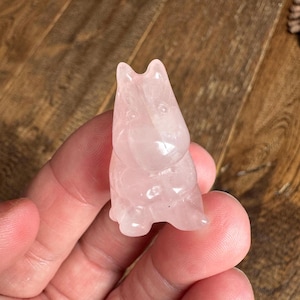 May include: A small, carved rose quartz figurine of a cat. The cat is sitting upright with its ears perked up. The figurine is a soft pink color and has a smooth, polished finish. The cat figurine is a decorative item.