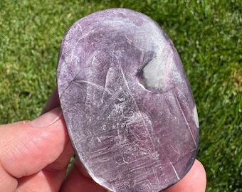 May include: A polished, oval-shaped purple stone with white inclusions and a textured surface. The stone is held in a person's hand against a backdrop of green grass. The stone's color ranges from light to dark purple.