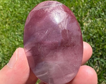 May include: A polished, oval-shaped stone with a deep burgundy color and subtle white veining. The stone is held in a hand against a backdrop of green grass, highlighting its smooth, reflective surface. A natural gemstone.