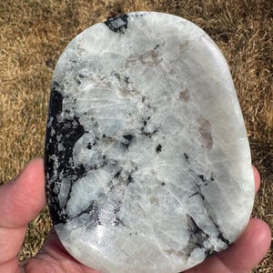 May include: A polished, oval-shaped stone with a marbled appearance. The stone features a mix of white, gray, and black colors, with dark veins and patches throughout. The surface is smooth and reflective, suggesting a high-quality finish.