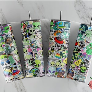May include: Four stainless steel tumblers with a white background and covered in colorful alien and UFO themed stickers. The tumblers have a silver lid and a straw.