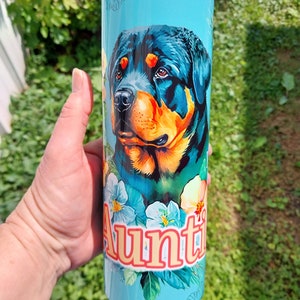 May include: A turquoise stainless steel tumbler with a colorful Rottweiler dog illustration and floral accents. The text "Auntie" is printed in orange on the tumbler.