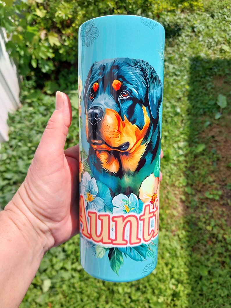 May include: A turquoise stainless steel tumbler with a colorful Rottweiler dog illustration and floral accents. The text "Auntie" is printed in orange on the tumbler.
