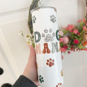 May include: White tumbler with a green, brown, and orange paw print design. The tumbler has the words "Dog Mom" written on it.