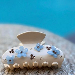 May include: A cream-colored hair claw clip featuring three light blue flower-shaped embellishments and several small, dark brown circular accents arranged across the curved surface of the clip, which has a standard spring-loaded jaw mechanism.