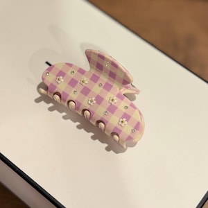 May include: A hair claw clip featuring a light purple and cream gingham check pattern. The surface is decorated with small white flower appliques with yellow centres and tiny clear rhinestones scattered across the checkered design.