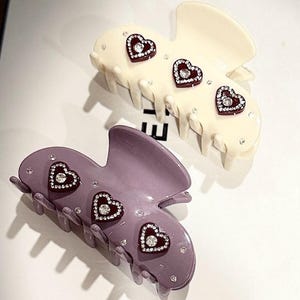 May include: Two claw hair clips, one cream and one mauve, each featuring three dark red heart-shaped embellishments with central clear rhinestones and additional scattered small clear rhinestones across the smooth, curved plastic surface.