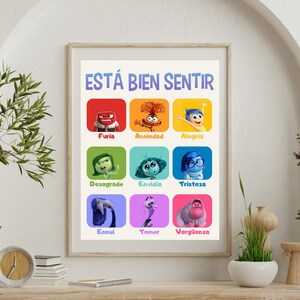 May include: A framed poster with the text "ESTÁ BIEN SENTIR" and illustrations of animated characters representing emotions. The characters are labeled with Spanish words like "Furia" and "Alegría". The poster is displayed on a wooden surface.