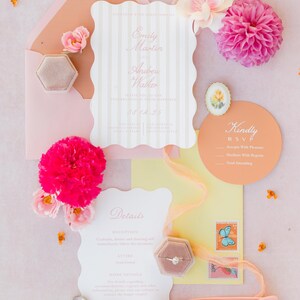 May include: A flat lay of wedding stationery in shades of pink and peach. Includes invitations, RSVP cards, a ring box, heart-shaped sunglasses, and flowers. The invitation reads "Emily Martin & Andrew Walker".