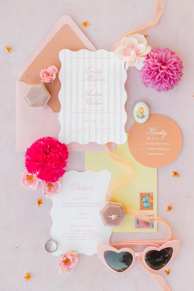 May include: A flat lay of wedding stationery in shades of pink and peach. Includes invitations, RSVP cards, a ring box, heart-shaped sunglasses, and flowers. The invitation reads "Emily Martin & Andrew Walker".