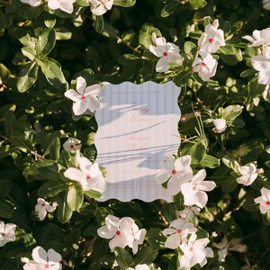 May include: A wedding invitation with a scalloped edge, featuring the names "Emily Martin" and "Andrew Hunter" in elegant script. The invitation is set against a backdrop of white flowers with red centers and lush green foliage.