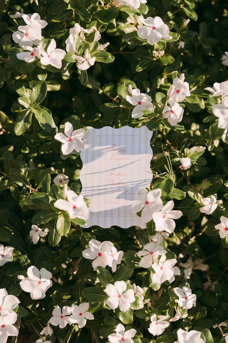 May include: A wedding invitation with a scalloped edge, featuring the names "Emily Martin" and "Andrew Hunter" in elegant script. The invitation is set against a backdrop of white flowers with red centers and lush green foliage.
