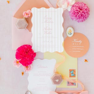 May include: A flat lay of wedding stationery, including invitations, RSVP cards, and details cards. The invitations have a scalloped edge and vertical stripes. Pink flowers, a ring box, and heart-shaped sunglasses are also present. The color palette is soft pink and peach.