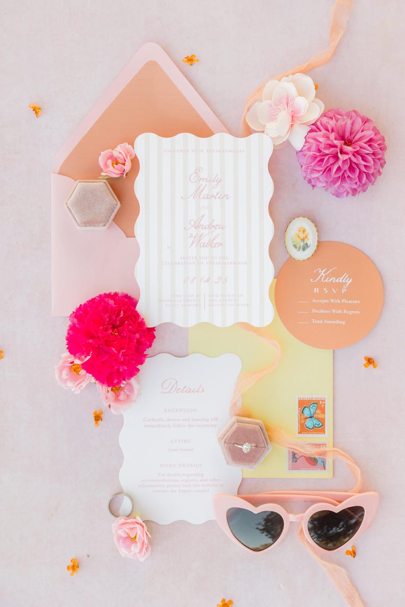 May include: A flat lay of wedding stationery, including invitations, RSVP cards, and details cards. The invitations have a scalloped edge and vertical stripes. Pink flowers, a ring box, and heart-shaped sunglasses are also present. The color palette is soft pink and peach.