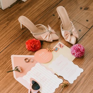 May include: A wedding flat lay featuring blush-colored high heels with bow accents and rhinestone straps. Wedding invitations, rings, sunglasses, and flowers are also arranged on a wooden surface, creating a romantic scene.