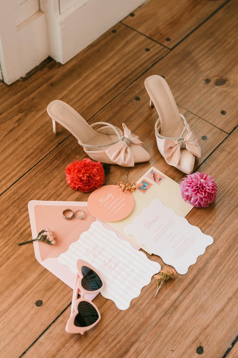 May include: A wedding flat lay featuring blush-colored high heels with bow accents and rhinestone straps. Wedding invitations, rings, sunglasses, and flowers are also arranged on a wooden surface, creating a romantic scene.