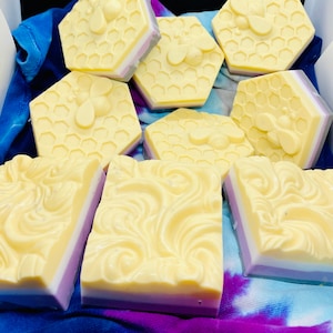 May include: A collection of handmade soap bars with a honeycomb design and bee accents. The soap bars are layered in shades of yellow, white, and purple.