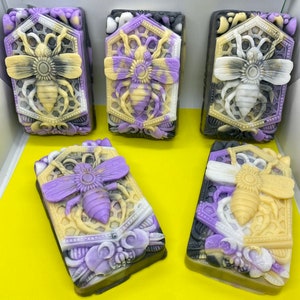 Può includere: Cinque saponette fatte a mano con un design a forma di ape e favo. Le saponette sono in tonalità di viola, nero, bianco e giallo.