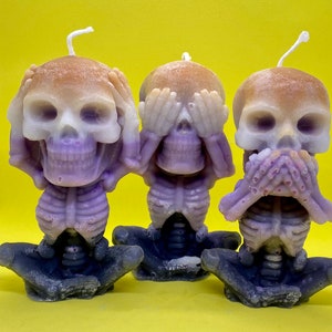 Peut inclure: Trois bougeoirs en forme de crâne, de couleur violet et blanc, chacun avec un geste de la main différent : l’un se couvrant les yeux, l’autre les oreilles et le dernier la bouche. Les bougies sont en cire et ont une finition mate.