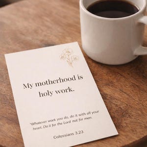 May include: A white ceramic mug filled with dark coffee sits on a wooden surface next to a card. The card reads "My motherhood is holy work" with a floral illustration and a Bible verse.